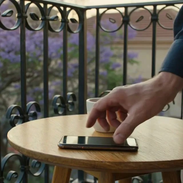 Una mano deja un celular en una mesita de balcón, con un jacarandá en flor detrás.